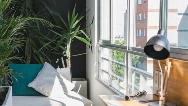 Una habitación moderna con un ventanal grande, un escritorio, una silla y un sofá. Hay plantas en macetas y una lámpara sobre el escritorio.