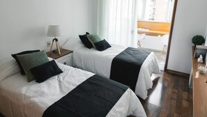 Habitación de hotel con dos camas, una mesita de noche con lámpara, una planta pequeña y vistas al exterior.