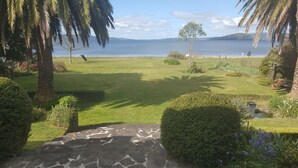 A well-maintained garden with a stone pathway leading to a grassy area, palm trees, and a view of a calm lake with distant hills. - Lake front luxury at your door step (Ngongotaha)