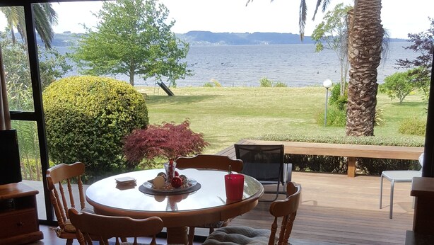 A dining area with a round table and chairs, a red candle, and a view of a lake and greenery. - Lake front luxury at your door step (Ngongotaha)