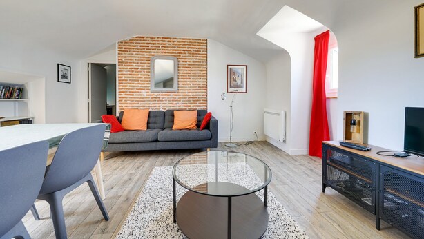 A modern living room with a brick accent wall, a glass coffee table, a grey sofa with orange cushions, and a red curtain. - The Heart of Amboise, three-room apartment, two bedrooms in the city center (Amboise)