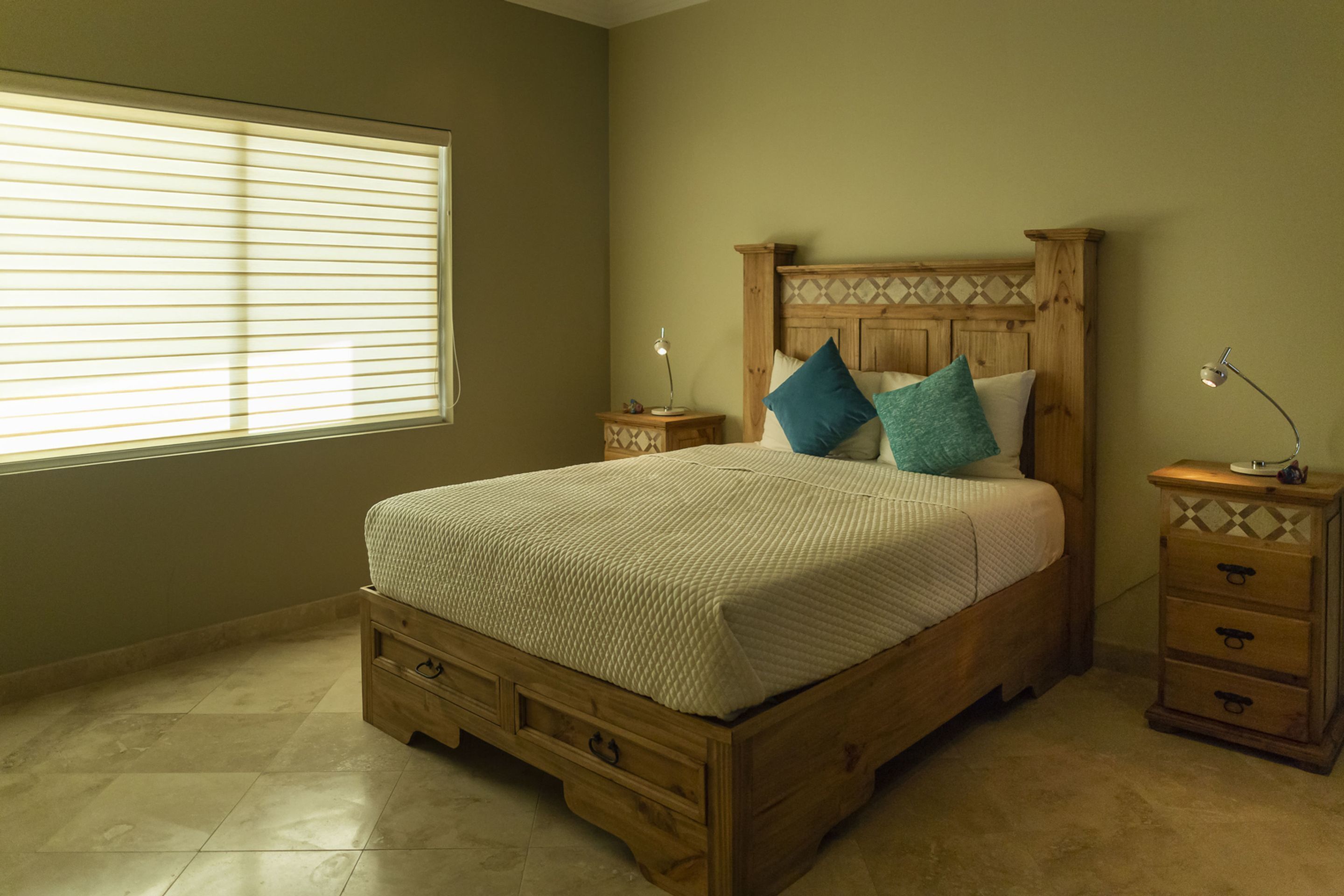 A bedroom with a wooden bed, two nightstands, a window with blinds, and a tiled floor.