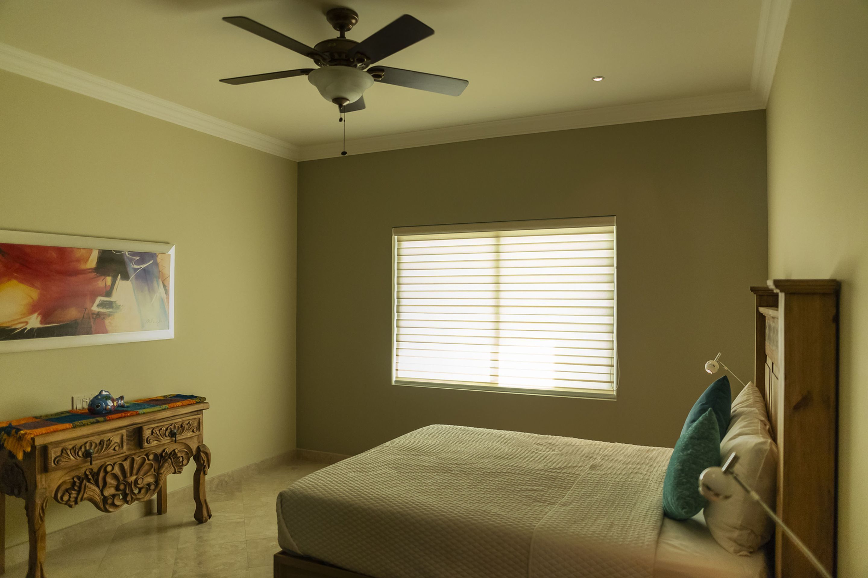 A bedroom with a bed, a ceiling fan, a window with blinds, a wooden dresser, and a painting on the wall.