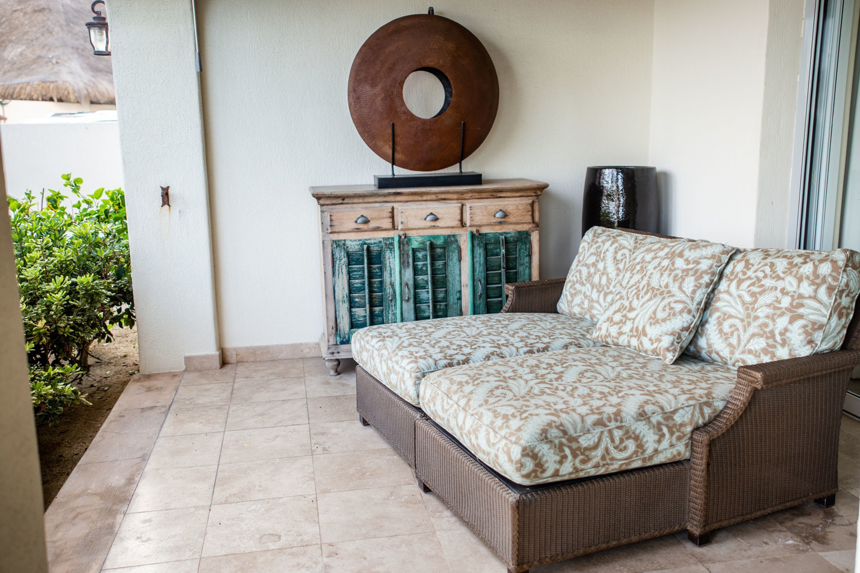 A wicker sofa with patterned cushions, a wooden cabinet with green shutters, and a large decorative plate on the wall.