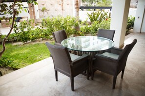 Een ronde glazen tafel met rieten stoelen in een terras, met uitzicht op een tuin en een gebouw op de achtergrond.
