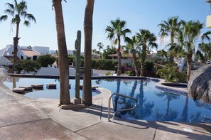 A pool area with palm trees, a cactus, and a building in the background. - La Costa 201 1 BR by Casago (San José del Cabo)