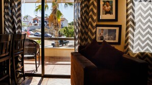 A room with a wooden sofa, a wicker chair, and a view of a street with palm trees and parked cars. - La Costa 201 1 BR by Casago (San José del Cabo)