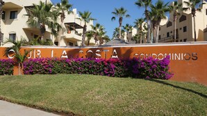 A building with a sign reading "CUB LA COSTA condominiums" surrounded by palm trees and purple flowers. - La Costa 201 1 BR by Casago (San José del Cabo)