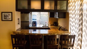 A kitchen with dark cabinets, a granite countertop, and a window with blinds. - La Costa 201 1 BR by Casago (San José del Cabo)