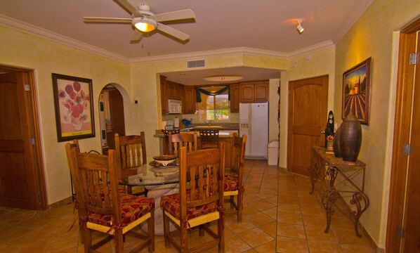 A dining area with wooden chairs and a glass table, a kitchen with wooden cabinets, a ceiling fan, and a tiled floor.