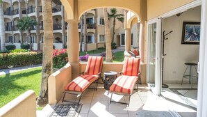 A balcony with two striped lounge chairs, a small round table, and a glass door leading to a pool area. - Las Olas A104 3 BR by Casago (San José del Cabo)