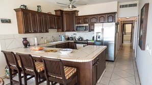 A kitchen with dark wooden cabinets, a large island with bar stools, a stainless steel refrigerator, and a microwave. - Las Olas A104 3 BR by Casago (San José del Cabo)
