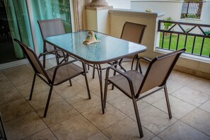 A glass-top table with four chairs, a small decorative item, and a view of a garden through a window with a metal railing. - Puerta Cabos Village G207 2BR by Casago (Cabo San Lucas)