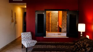A hotel room with a red wall, a bed with a patterned bedspread, a chair with a floral design, a bathroom visible through a window, and a lamp on the nightstand. - Puerta Cabos Village G207 2BR by Casago (Cabo San Lucas)
