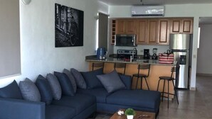 A modern living room with a dark blue sectional sofa, a wooden coffee table, and a ceiling fan. - Peninsula 106 2 BR by Casago (San José del Cabo)