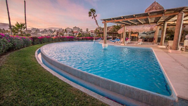Una piscina con borde curvo, rodeada de césped y una estructura cubierta de paja con sillones de descanso.