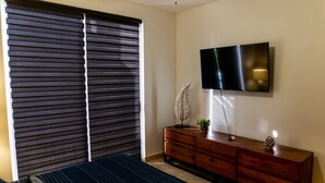 Un dormitorio con cama, cómoda, televisor montado en la pared y ventana con persianas.