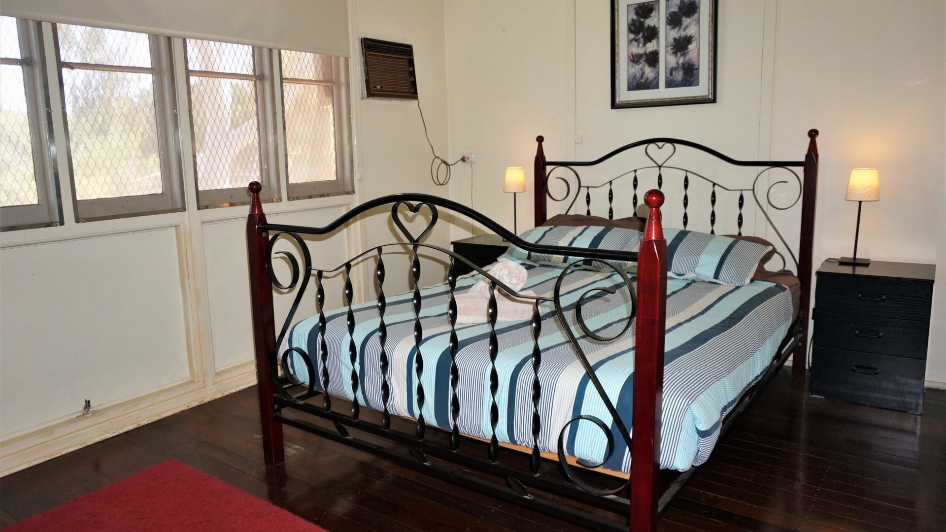 A bed with a metal frame, a red accent, and striped bedding. There is a bedside table with a lamp, a framed picture on the wall, and a window with a view.