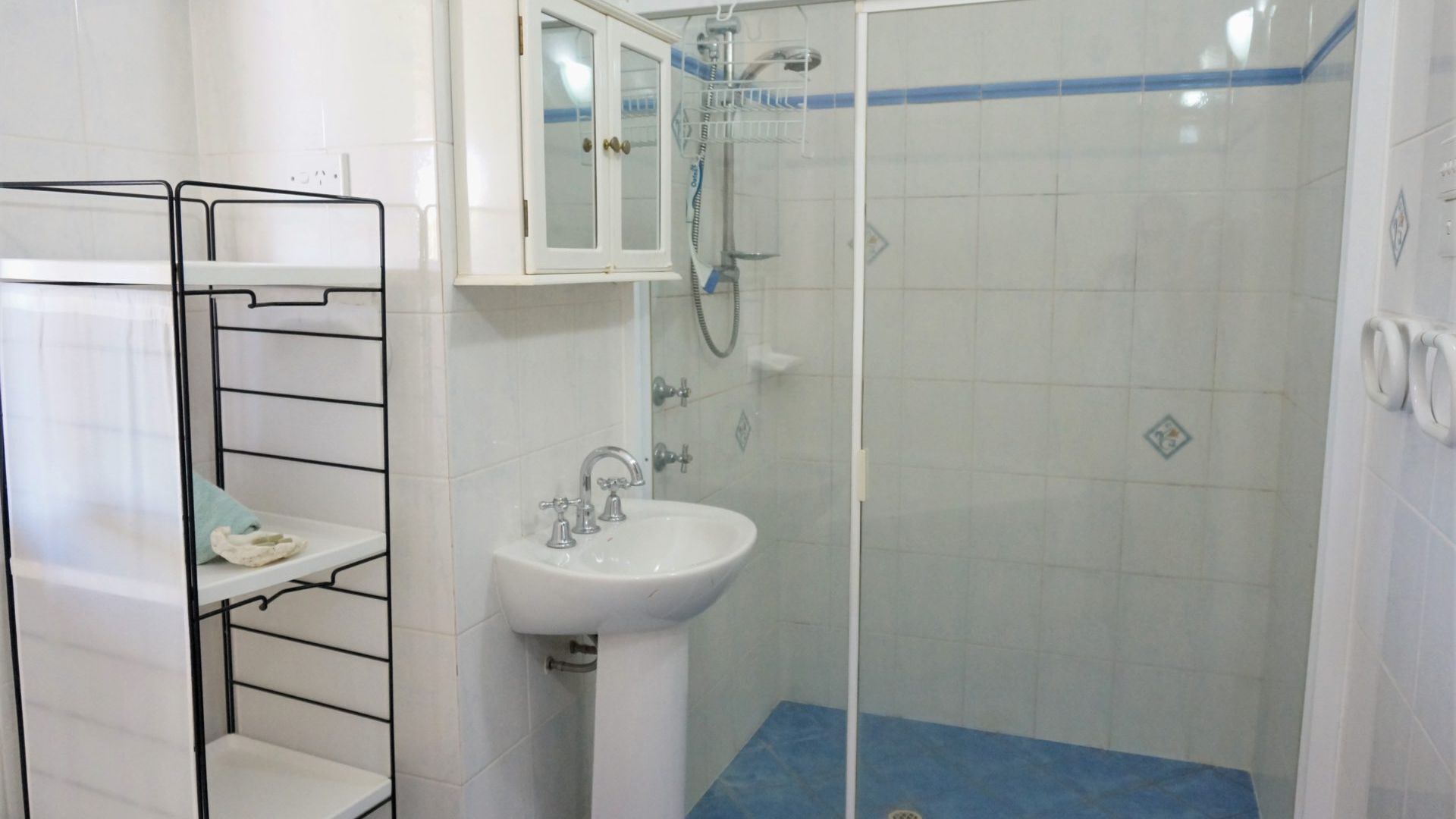 Un baño con ducha envuelta en vidrio, un lavabo blanco y una estantería de metal.