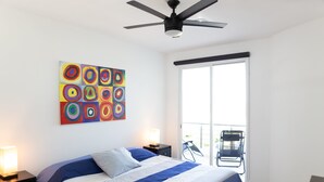 Un dormitorio con cama, ventilador de techo, mesita de noche con lámpara, un cuadro abstracto de colores y una puerta corrediza de vidrio que da a un espacio exterior.