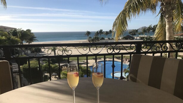 A balcony with two glasses of a light-colored beverage, overlooking a pool and beach. - Las Mananitas E3301 2 BR by Casago (San José del Cabo)