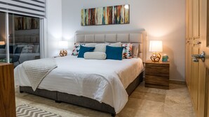 A bedroom with a large bed, a headboard, two bedside tables, a wall-mounted artwork, and a sliding door. - Peninsula 103 2 BR by Casago (San José del Cabo)