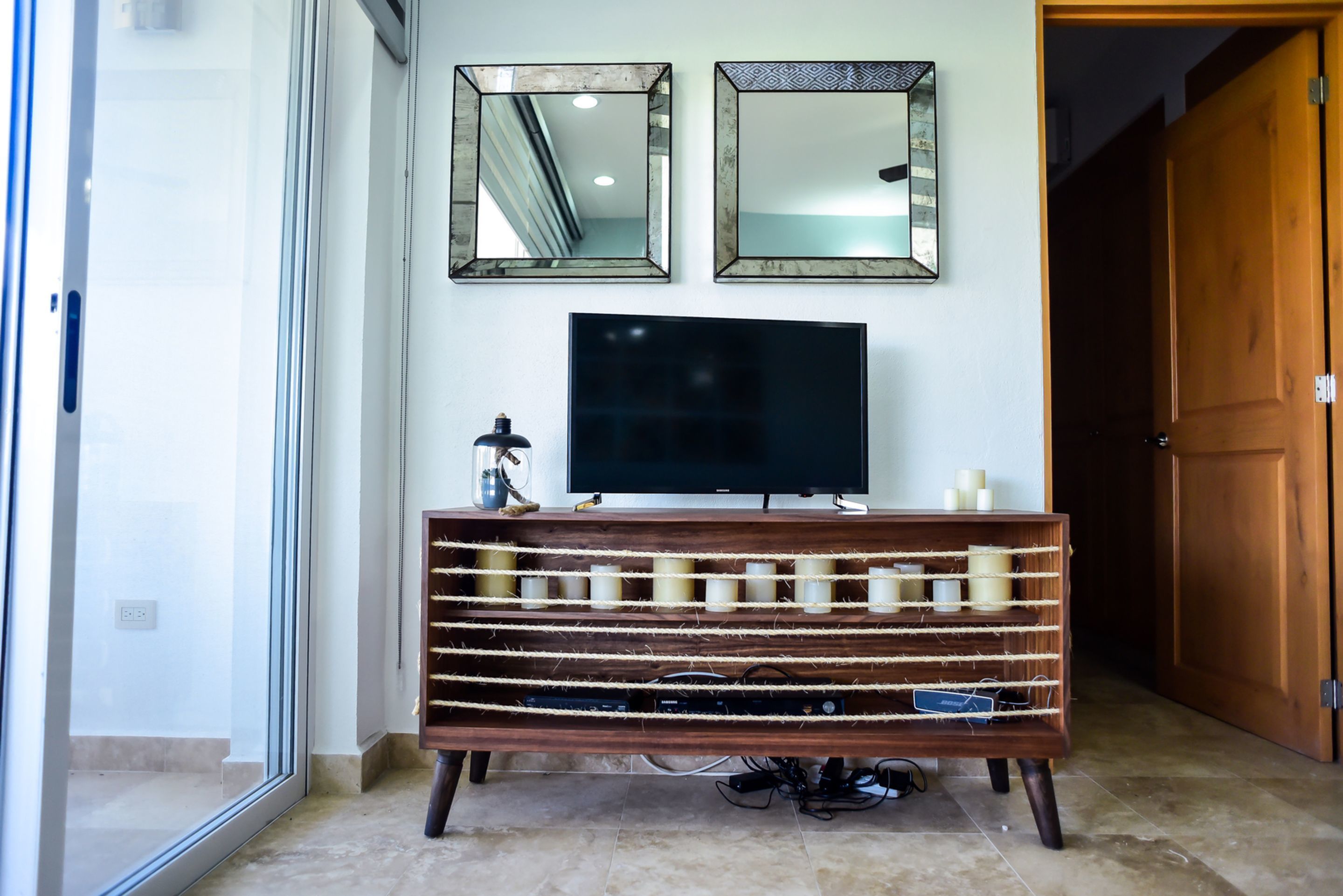 Una habitación con una televisión sobre un mueble de madera, dos espejos en la pared y una puerta a la derecha.