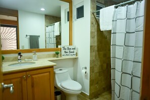 Un baño con inodoro, lavamanos y cortina de ducha. Hay un cartel en la pared que dice 'El Sol y la Arena y un Trago en mi Mano'.