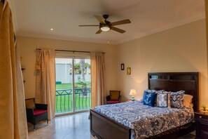Une chambre à coucher comprenant un lit, un ventilateur de plafond, une table de chevet, une chaise et une vue sur un espace extérieur verdoyant.