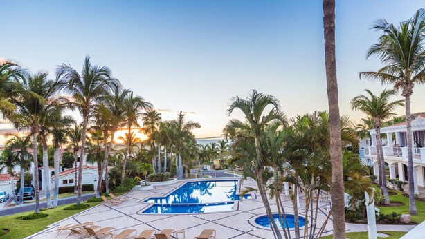 A pool area with lounge chairs, palm trees, and a building in the background. - Villa De Oro 106 3 BR by Casago (Cabo San Lucas)