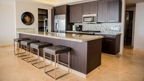 A modern kitchen with dark wood cabinets, stainless steel appliances, and a marble countertop. There are four bar stools with cushioned seats. - Cascadas de Pedregal 711 3 BR by Casago (Cabo San Lucas)
