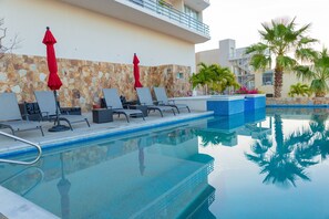 A swimming pool with lounge chairs and red umbrellas, surrounded by palm trees and a stone wall. - Cascadas de Pedregal 711 3 BR by Casago (Cabo San Lucas)