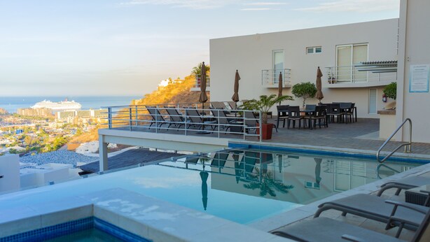 A rooftop infinity pool with lounge chairs and tables, overlooking a cityscape and the ocean. - Cascadas de Pedregal 711 3 BR by Casago (Cabo San Lucas)