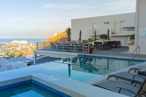 A rooftop infinity pool with lounge chairs and tables, overlooking a cityscape and the ocean. - Cascadas de Pedregal 711 3 BR by Casago (Cabo San Lucas)