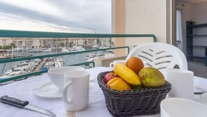 Un balcon avec une table dressée pour deux personnes, un panier en osier rempli de fruits et une vue sur un port de plaisance.