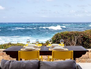 A dining table set with plates, glasses, and cutlery, overlooking a rocky coastline with the ocean in the background.