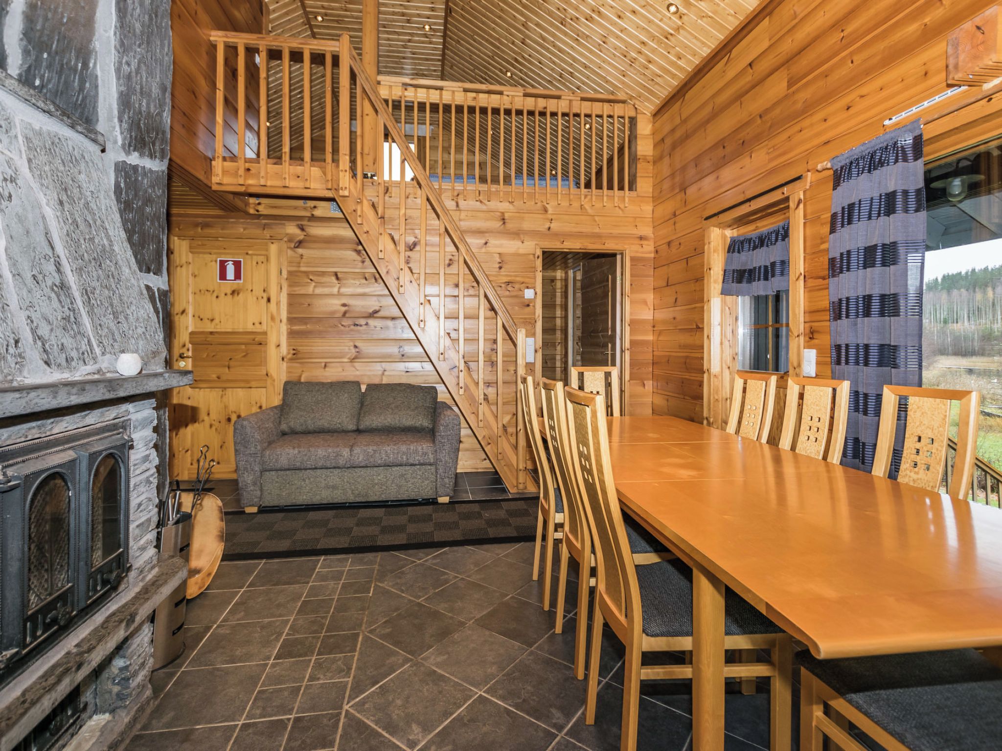 Interior de una cabaña de madera con chimenea, un sofá, una mesa de comedor con sillas y una escalera.