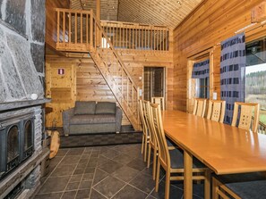 A wooden cabin interior with a fireplace, a sofa, a dining table with chairs, and a staircase. - Nuppulanranta 14 by Interhome (Kuhmoinen)