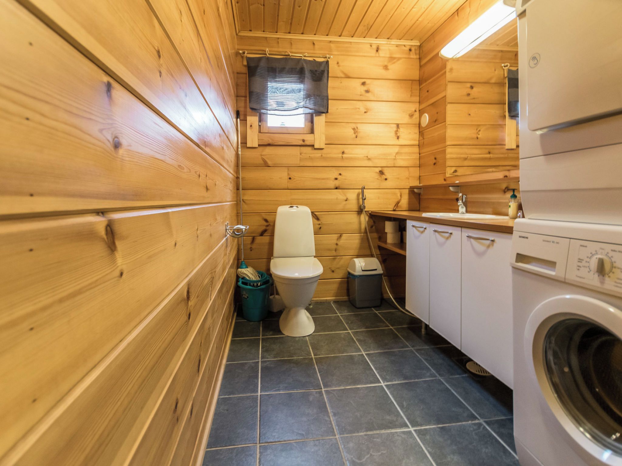 Et lille badeværelse i træ med toilet, vaskemaskine og et vindue.