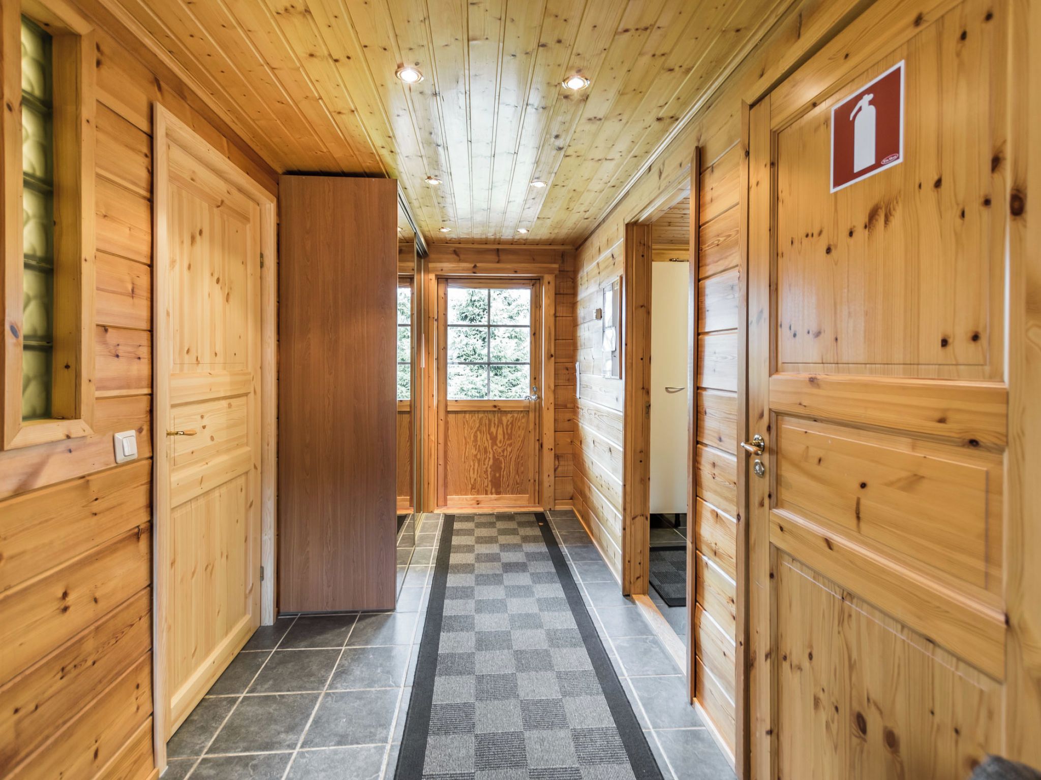 Un corridoio in legno con porte su entrambi i lati, un tappeto a scacchi sul pavimento e una finestra in fondo.