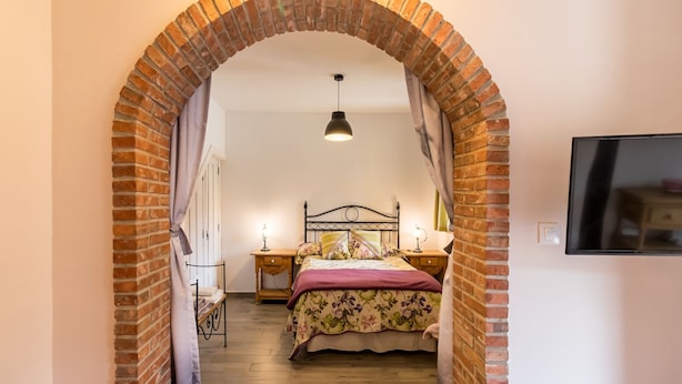Ein Schlafzimmer mit Ziegelbogen, einem Bett mit floralem Bettzeug, einem Nachttisch mit Lampe und einem an der Wand montierten Fernseher.