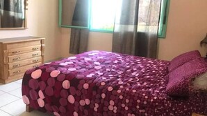 A bedroom with a bed covered in a patterned bedspread, a wooden dresser, a mirror, and a window with curtains. - Residence Cite Jardin 4065 (Martil)
