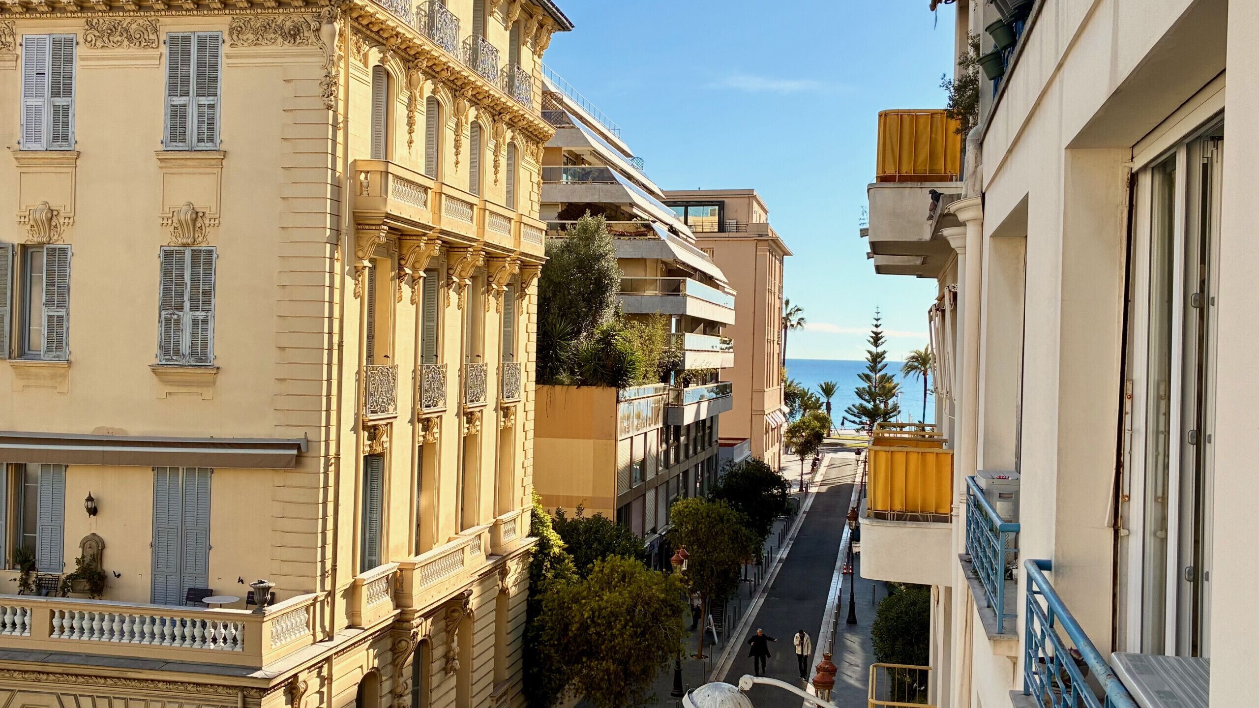 A narrow street with buildings on either side, featuring balconies and a view of the sea.