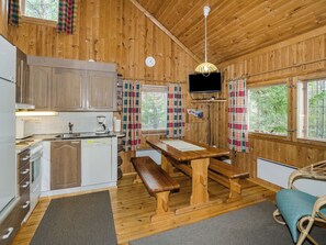 A cabin interior with wooden walls, a kitchen area, a dining table with benches, a television mounted on the wall, and a window with curtains. - Kanerva by Interhome (Juuka)