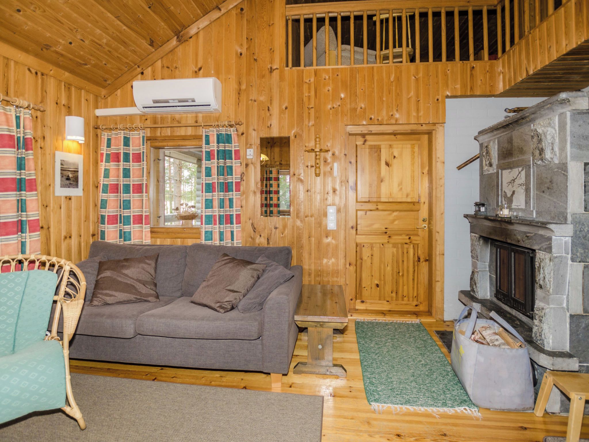 L’intérieur d’un chalet chaleureux, avec un canapé gris, des murs en bois, une cheminée et une fenêtre ornée de rideaux à carreaux.