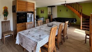 A spacious dining area with a long table, wooden chairs, and a patterned tablecloth. There is a kitchen with wooden cabinets and a built-in oven. A staircase with a wooden railing leads to an upper level.