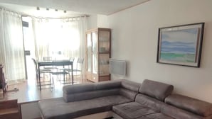 A living room with a sectional sofa, a glass coffee table, a dining area with a table and chairs, and a cabinet with glass doors. - Beautiful comfortable apartment at the foot of the slopes with internet (Isola 2000)
