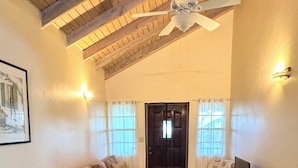 Ein Wohnzimmer mit hoher Decke, einem Deckenventilator, einem braunen Sofa, einem blauen Sofa, einem Fernseher auf einem Holzboden und einer Tür mit Vorhängen.