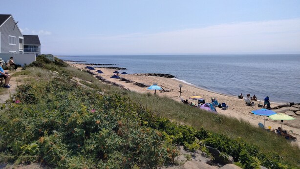 Ein Strand mit Liegestühlen und Sonnenschirmen, ein Haus links und das Meer im Hintergrund.