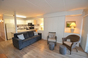 A living room with a grey sofa, two wicker chairs, a small ottoman, and a kitchen in the background.